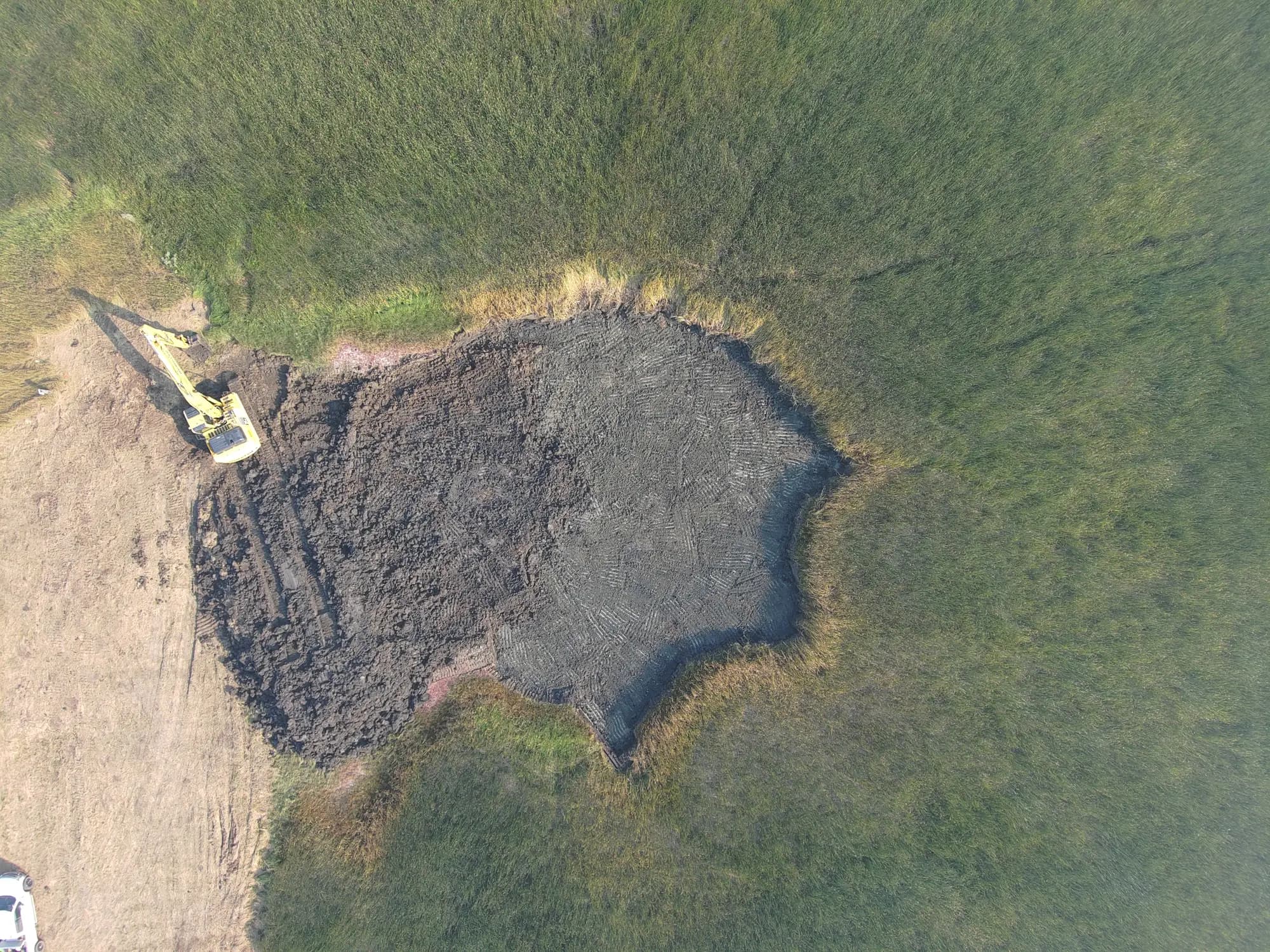 Aerial view of an excavator opening up the impacted ground at the edge of a pond, thick grass all around.